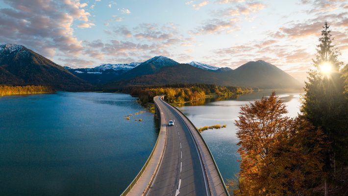 Car crossing a scenic bridge over a lake with mountains in the background, symbolizing navigating the path through 2025’s evolving regulatory landscape for sustainability.