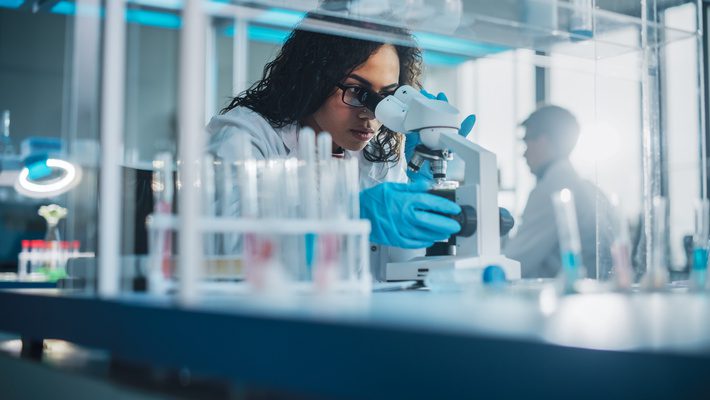 A scientist in a laboratory examines a sample under a microscope, surrounded by test tubes.